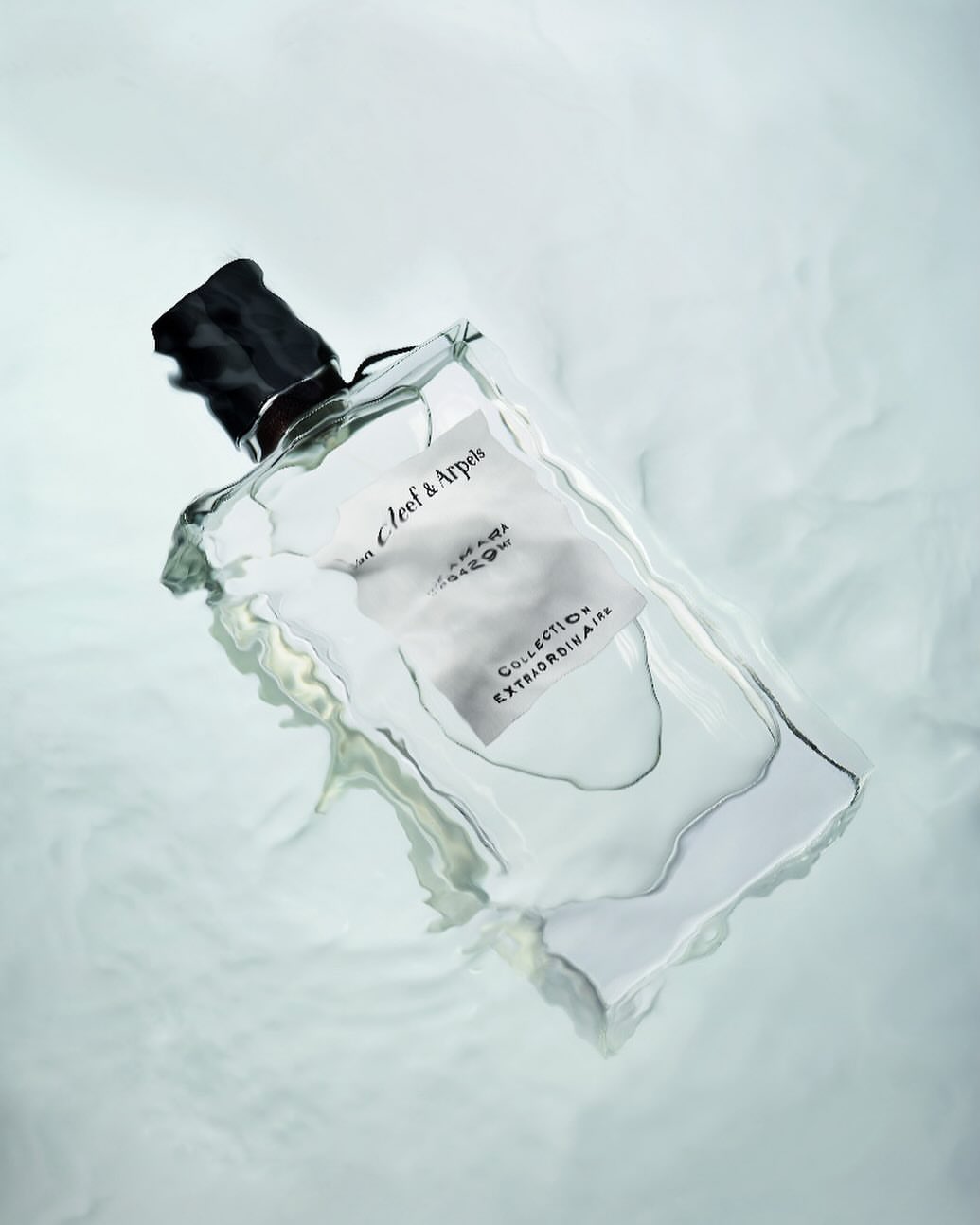 A glass perfume bottle with a black cap and a white label floats in a blurry, light-blue, dreamy background.
