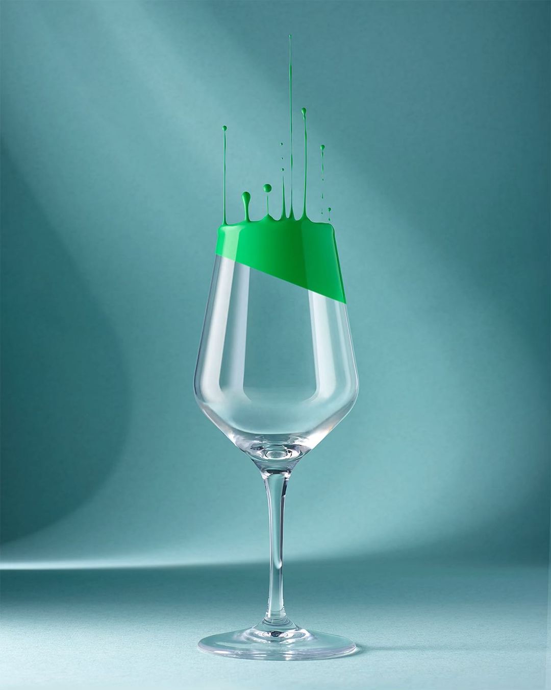 A clear wine glass is partially filled with bright green liquid, some of which drips down the glass against a serene teal background.