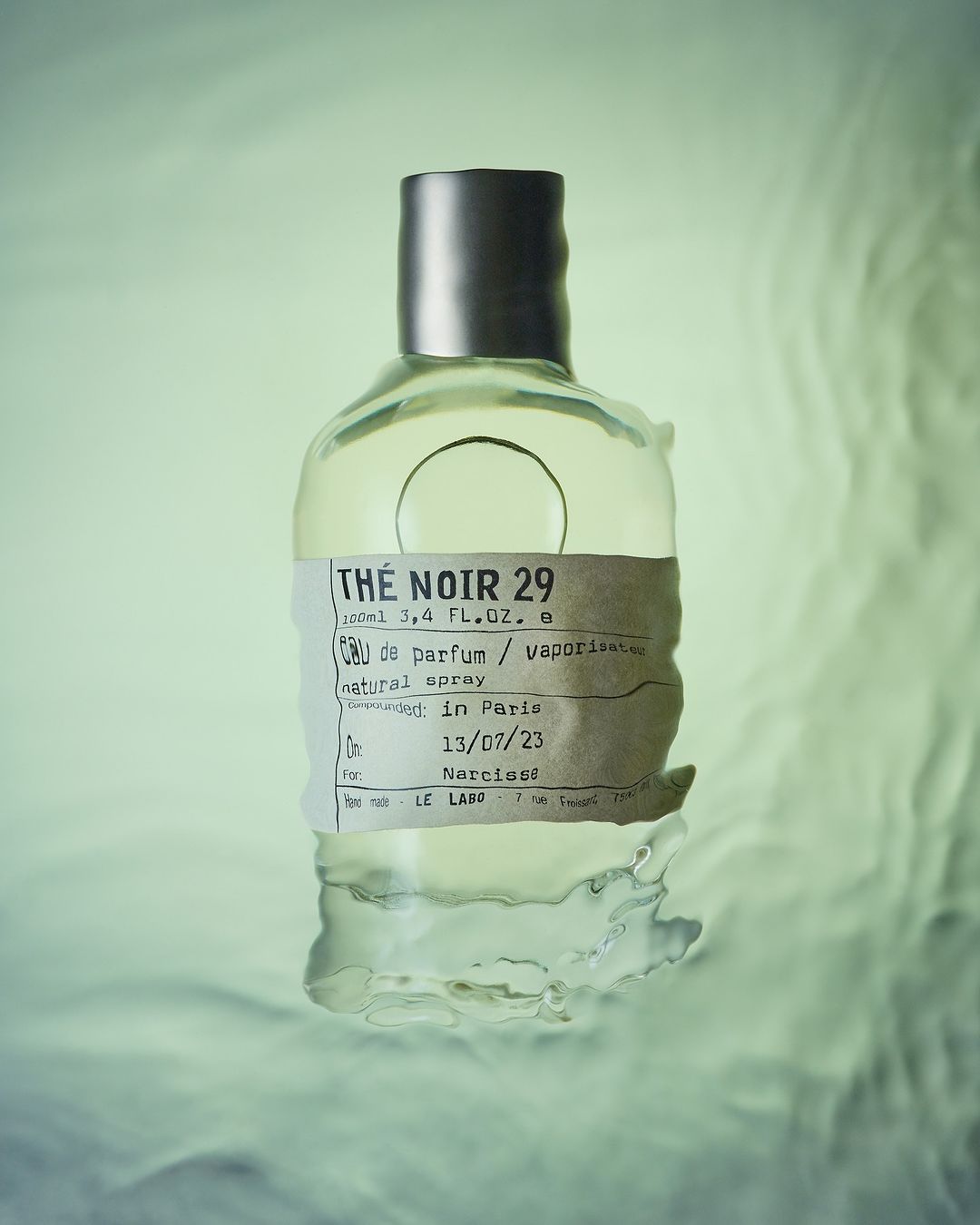 Close-up studio shot of a clear Le Labo Thé Noir 29 perfume bottle with silver cap and label, set against a soft green background.