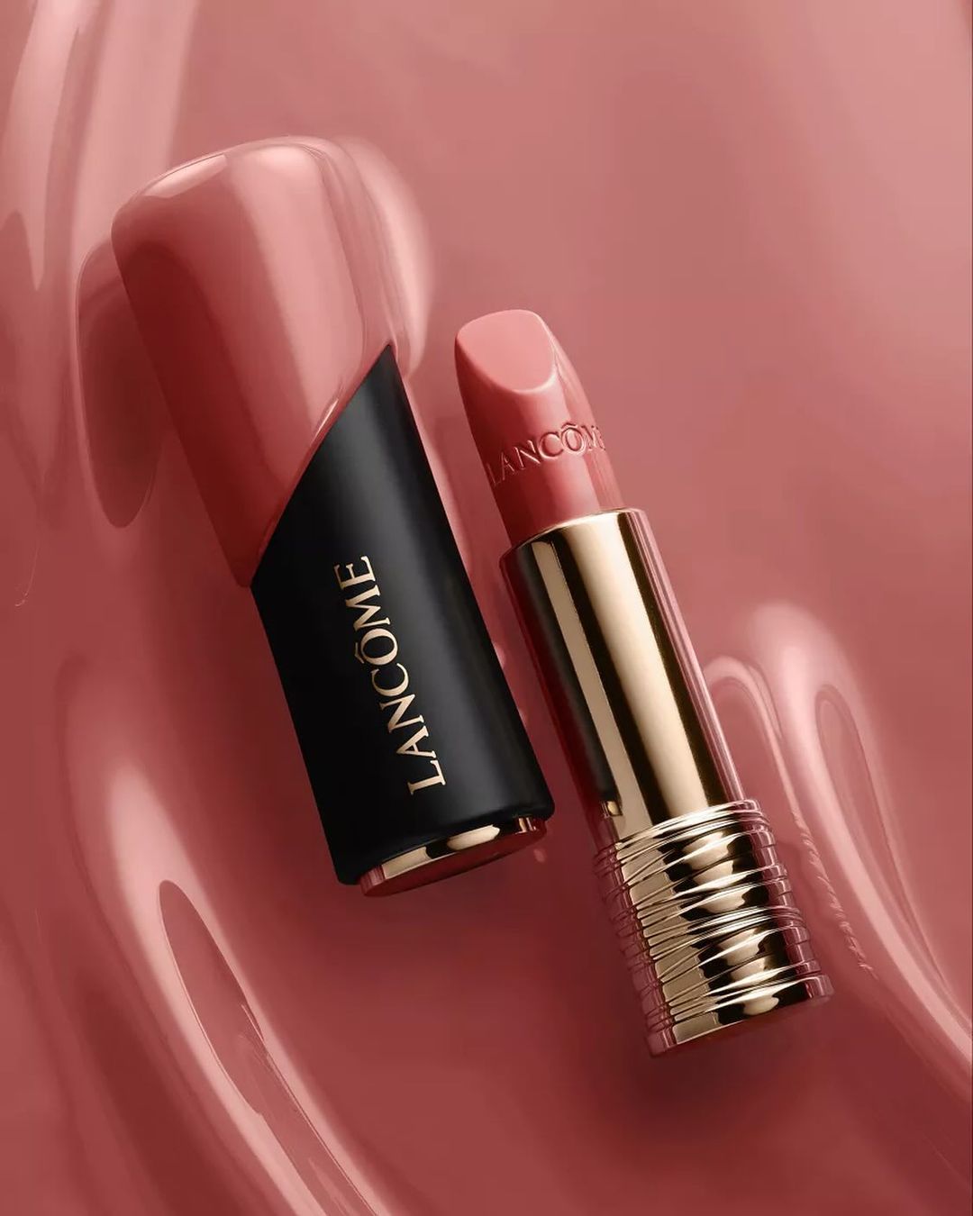 Product shot of Lancôme lipstick with black and gold case, next to its cap, on a glossy pink surface in a studio setting.