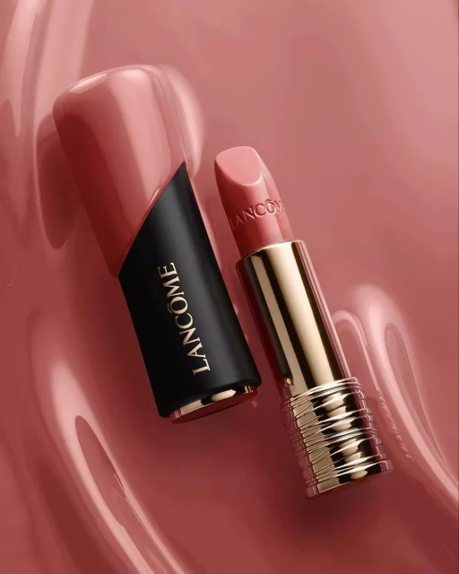 Product shot of Lancôme lipstick with black and gold case, next to its cap, on a glossy pink surface in a studio setting.