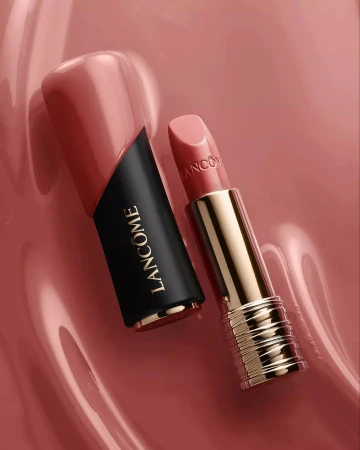 Product shot of Lancôme lipstick with black and gold case, next to its cap, on a glossy pink surface in a studio setting.