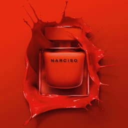 A square product photograph of a red perfume bottle surrounded by a dynamic red liquid splash on a matching red background.