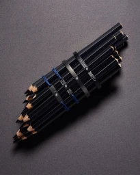 A bundle of black pencils arranged diagonally with jewellery bracelets resting across them on textured grey