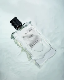 A rectangular glass perfume bottle submerged in rippling water with soft light filtering through.