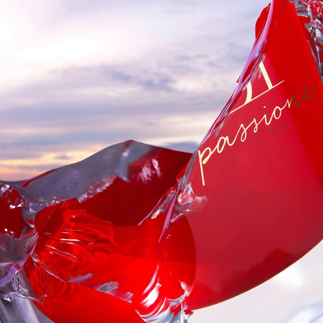 Abstract image featuring a red, liquid-like object with gold text against a soft, cloudy sky, creating a surreal and dynamic composition.
