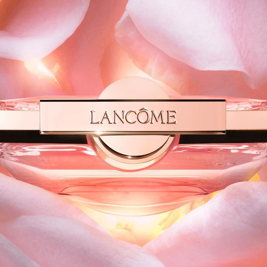 Close-up of a Lancôme perfume bottle with a gold-tone cap, centered on a background of soft-focus pink and white rose petals.