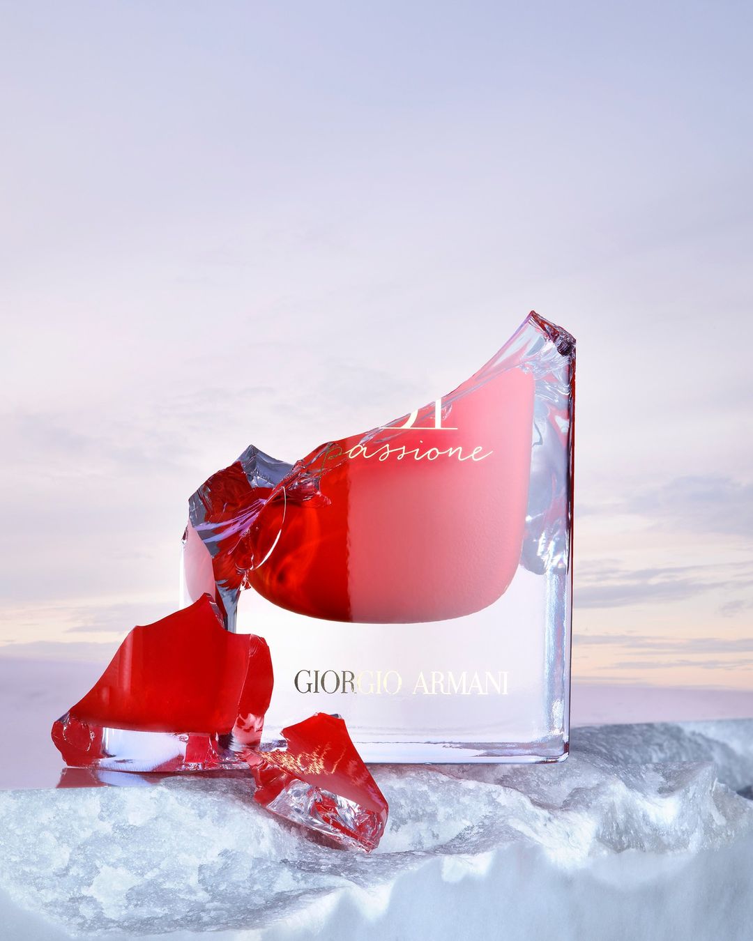 Giorgio Armani perfume bottle on reflective surface, shards of glass, red accents, against a soft sky-blue gradient background.