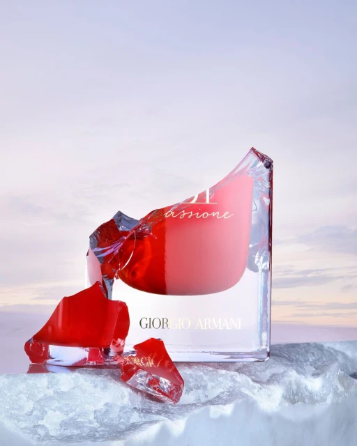 Giorgio Armani perfume bottle on reflective surface, shards of glass, red accents, against a soft sky-blue gradient background.