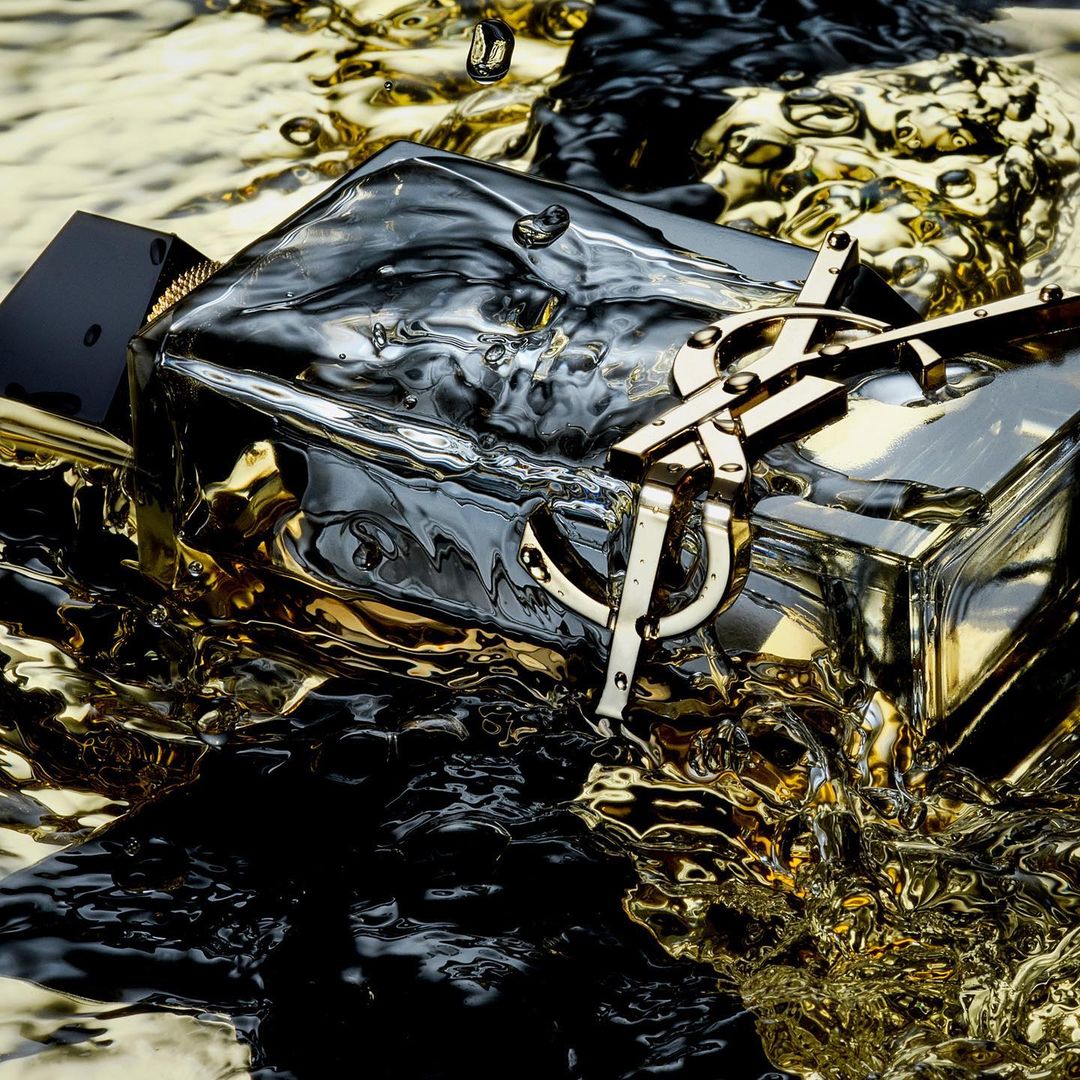 Close-up of a black and gold perfume bottle with the YSL logo, surrounded by liquid and water droplets, creating a dynamic splash effect.