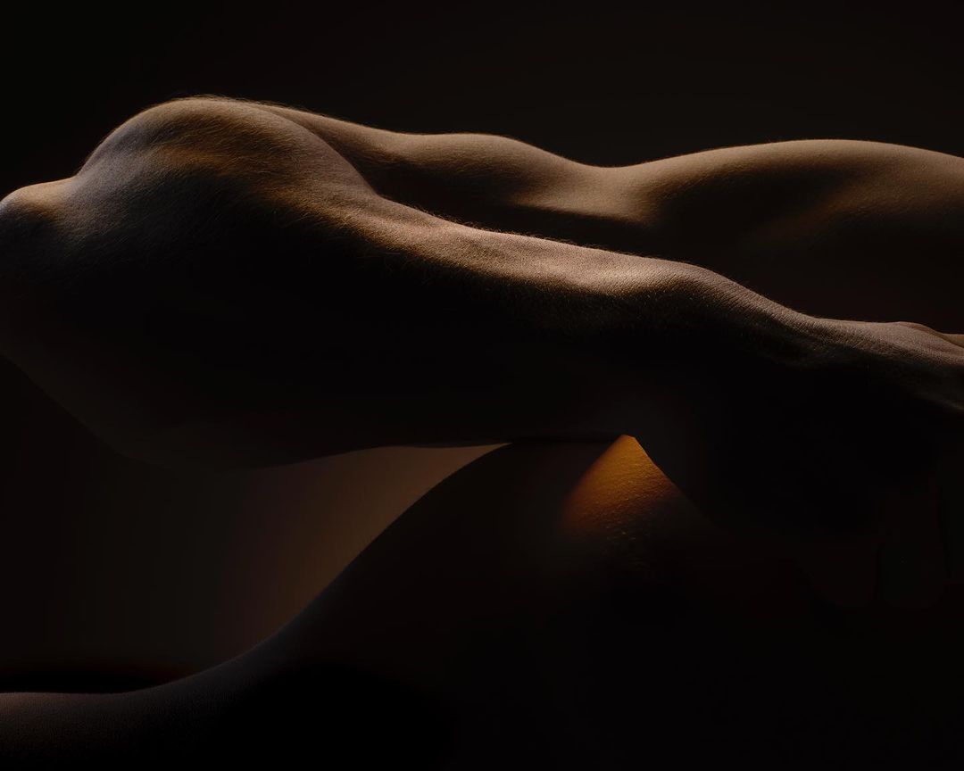 Nude human body in low-key lighting, showcasing muscular definition and curves in a photo studio setting against a dark background.