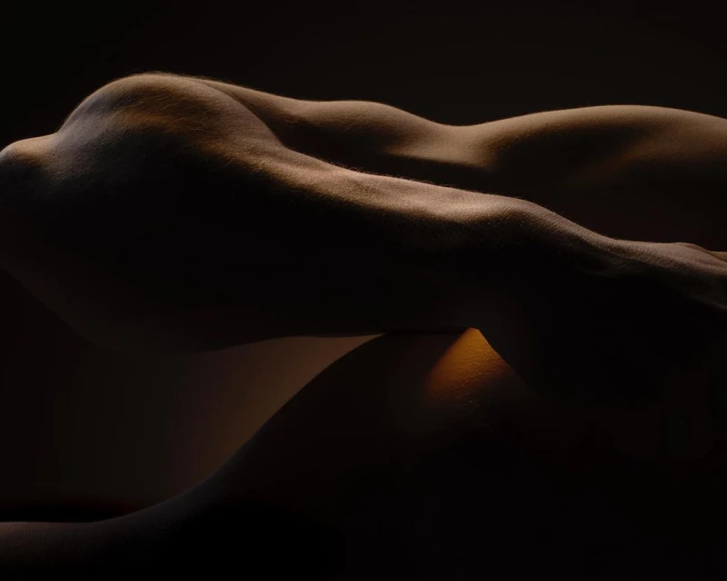 Nude human body in low-key lighting, showcasing muscular definition and curves in a photo studio setting against a dark background.