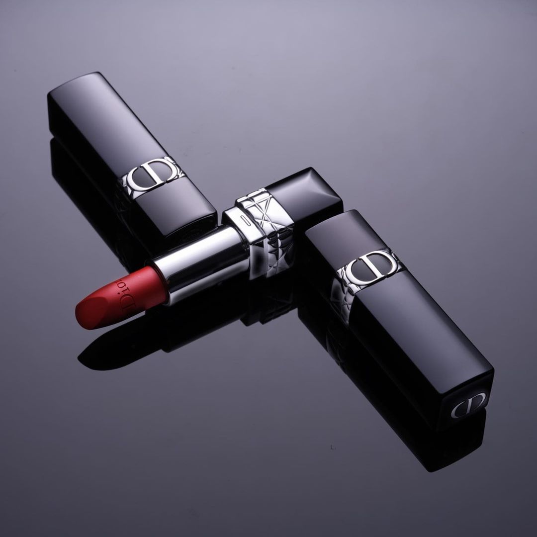 Still life photograph of three Dior brand lipsticks with red color visible, displayed on a glossy black surface in studio lighting.
