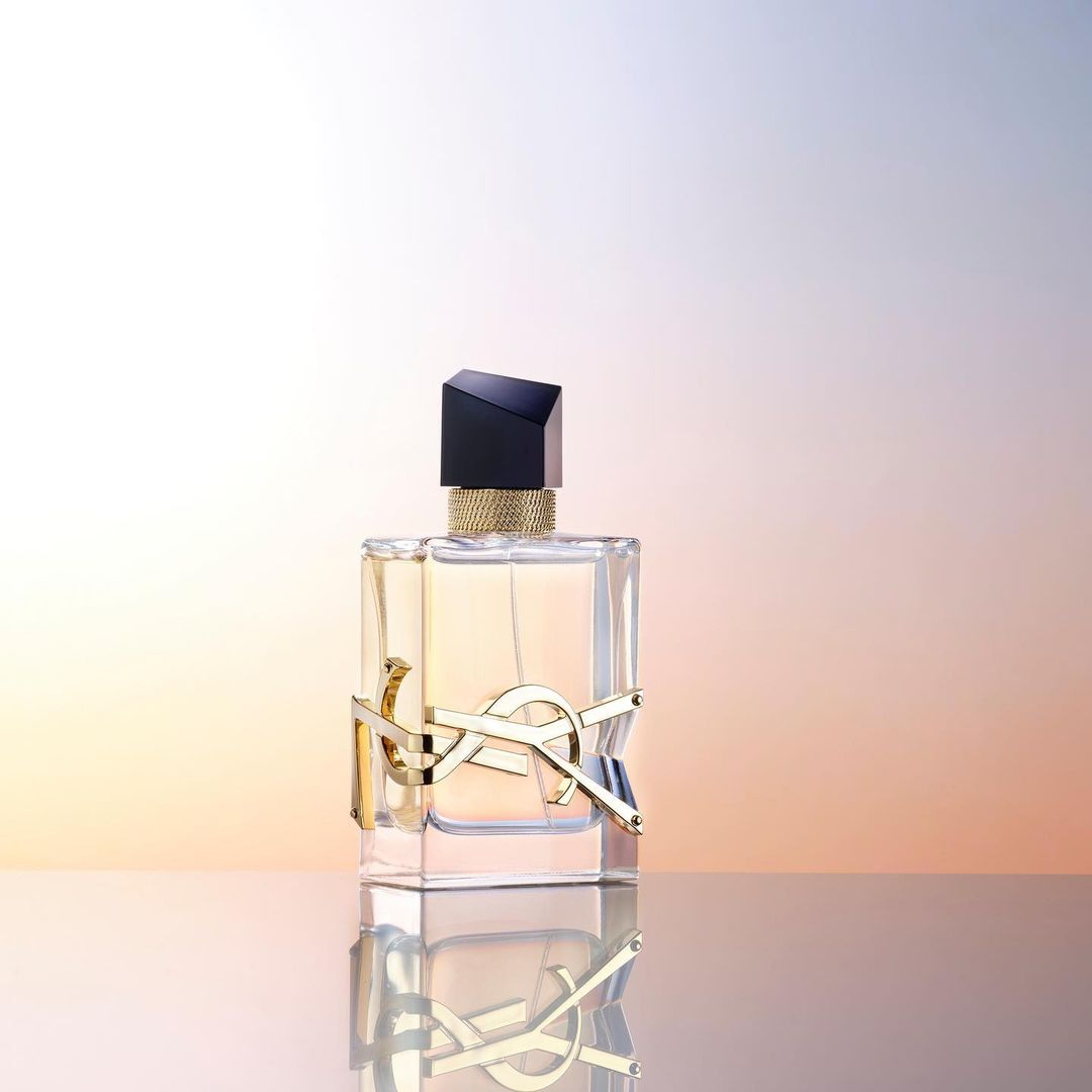 A glass perfume bottle with gold logo detail on a reflective surface against a gradient background with pink, white, and yellow hues.