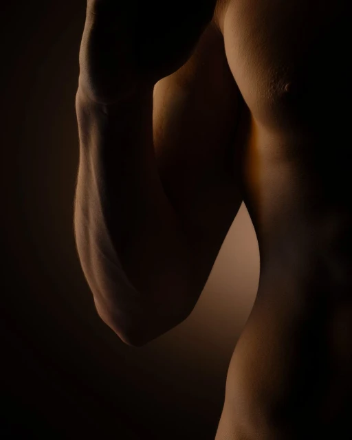 Torso and arm of a muscular adult male are visible against a dark background, highlighting muscle definition and form in a close-up shot.
