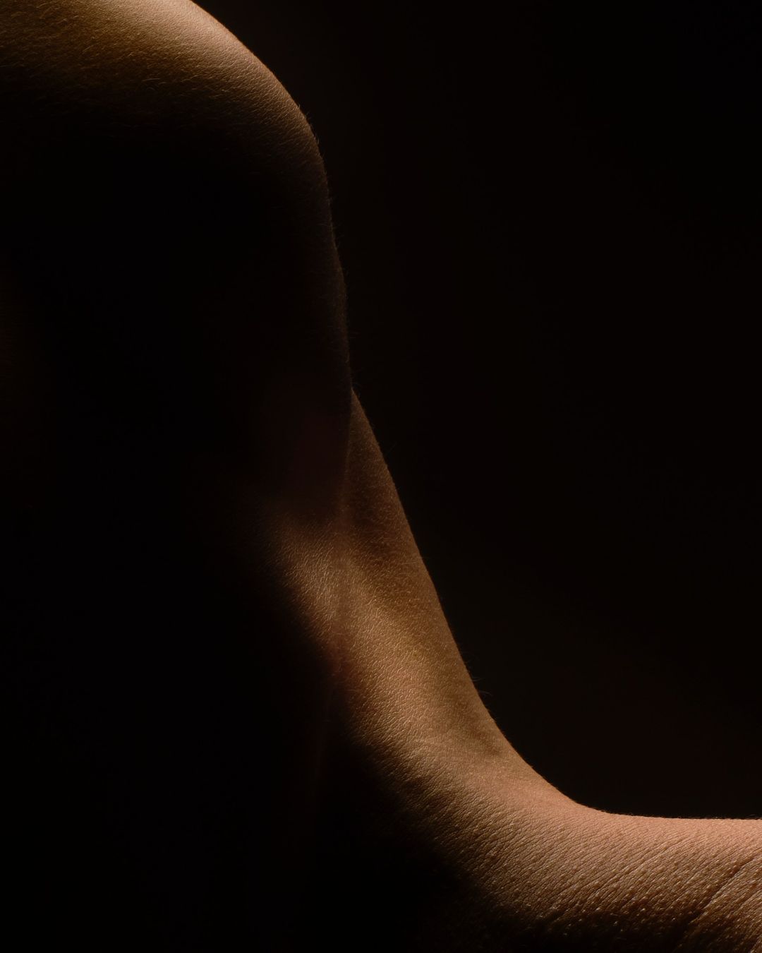 Close-up of a human arm against a dark background, showcasing skin texture and subtle shadows. Lighting highlights the form.