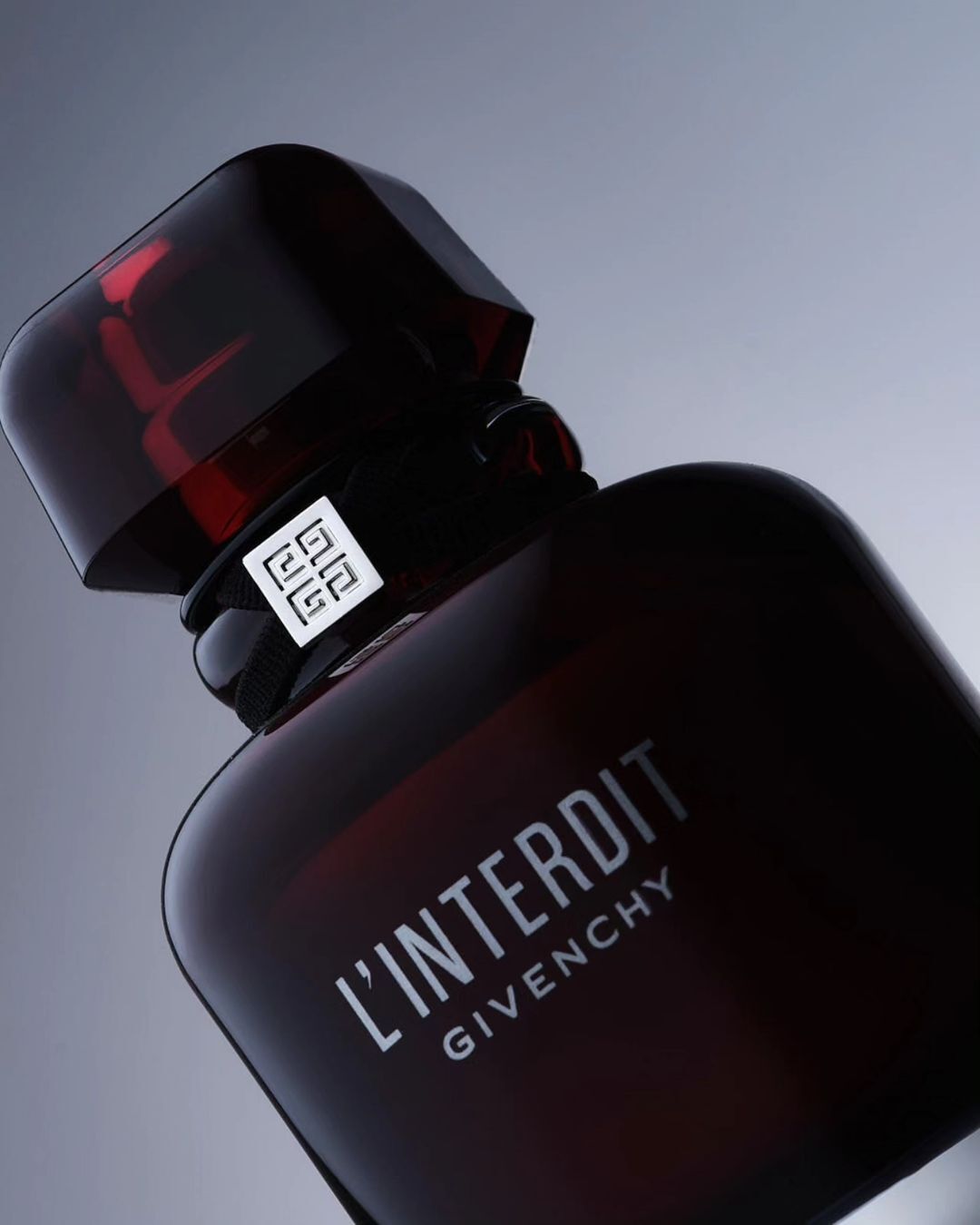Close-up studio shot of a bottle of L'Interdit Givenchy perfume with a dark red hue and translucent cap, against a grey gradient backdrop.