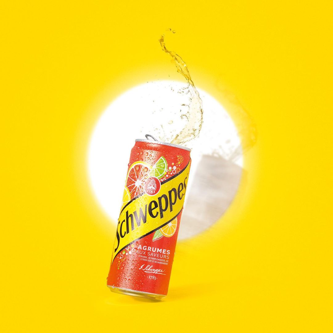 Red and yellow Schweppes can with splashing liquid against a bright yellow backdrop with a soft white circular gradient.