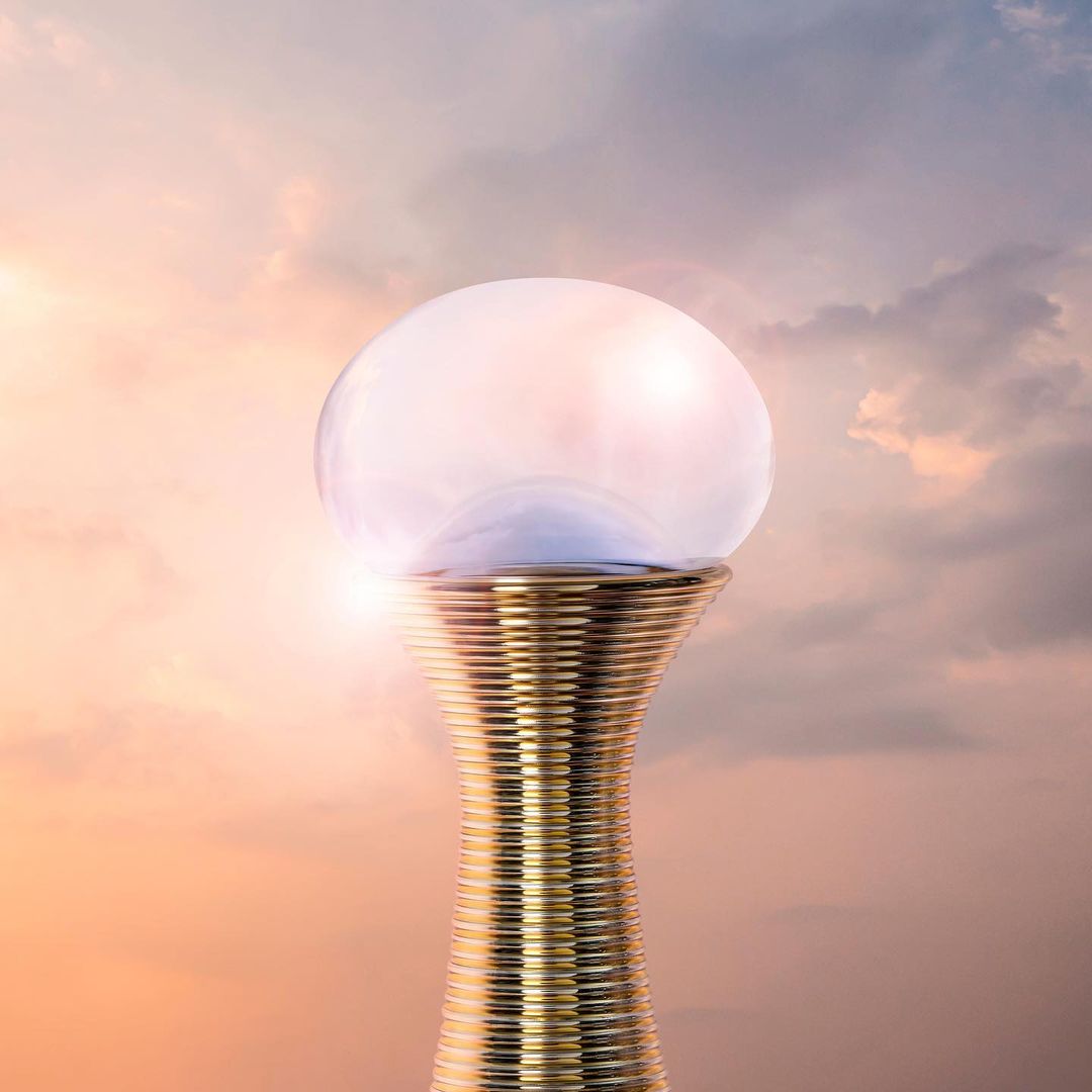 Gilded perfume bottle with clear spherical cap on a cloudy, twilight sky backdrop. Beauty product against a dramatic evening sky.