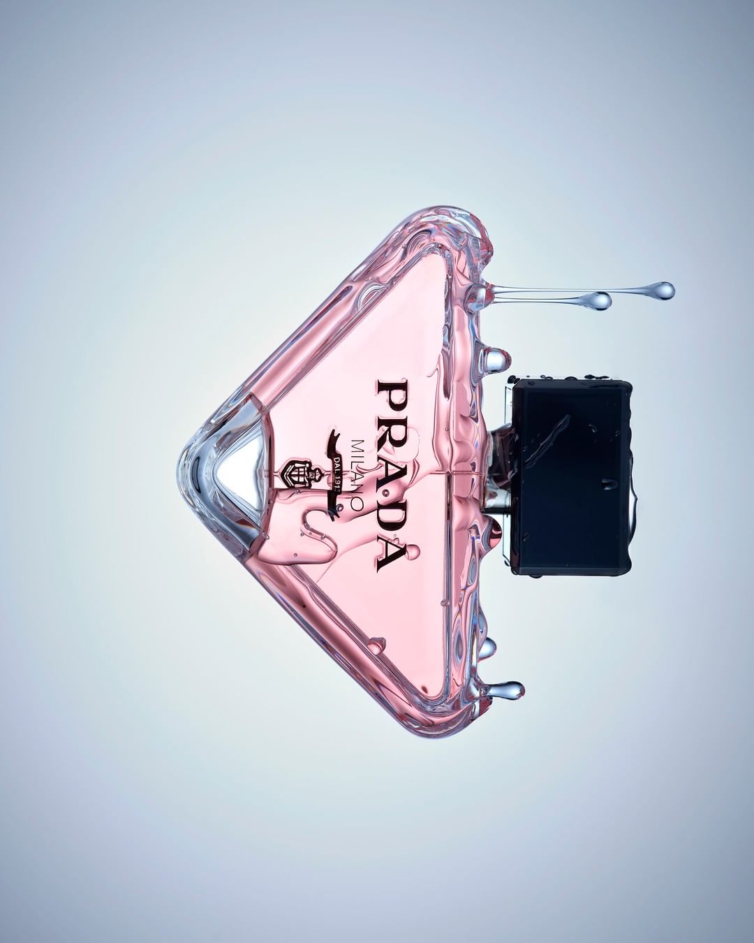 Prada perfume bottle in a triangular shape with a pink liquid inside and a black atomizer is surrounded by splashing water.