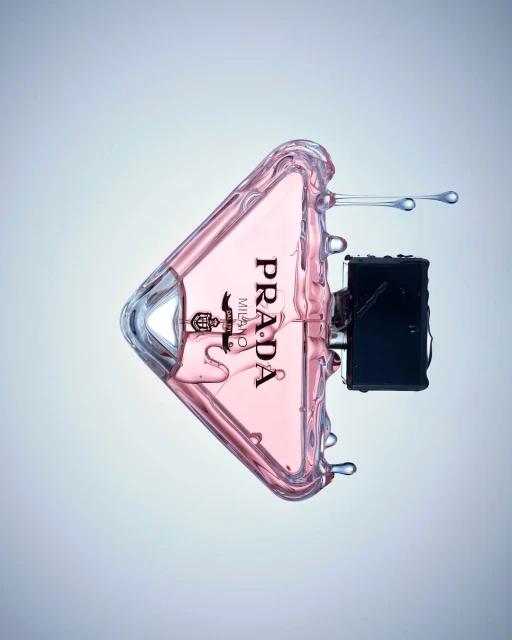 Prada perfume bottle in a triangular shape with a pink liquid inside and a black atomizer is surrounded by splashing water.