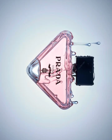 Prada perfume bottle in a triangular shape with a pink liquid inside and a black atomizer is surrounded by splashing water.