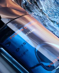 Close-up product photograph of a dark blue perfume bottle with splashing water across the top edge against a warm metallic background