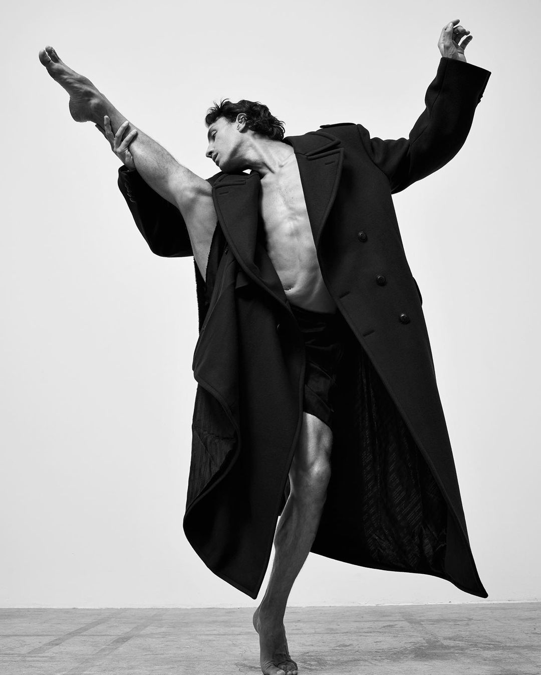 Black and white studio shot of male ballet dancer in a black coat performing a leg extension. Artistic dance and fashion concept.