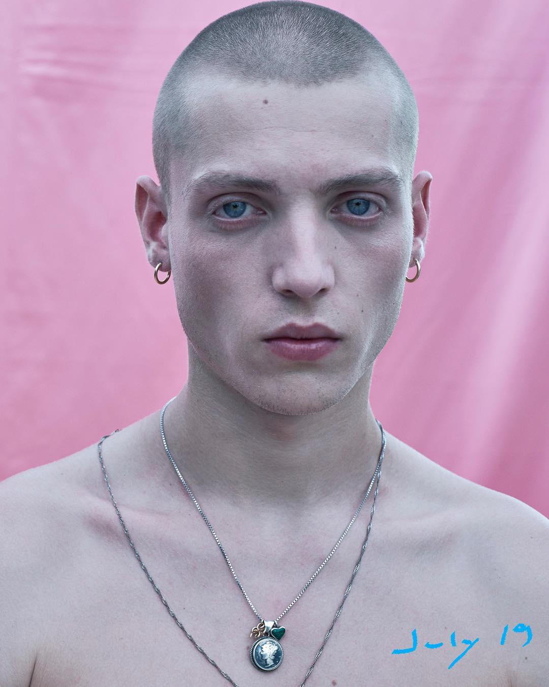 Portrait of a young adult male with shaved head against a pink background, wearing hoop earrings and pendant necklaces.