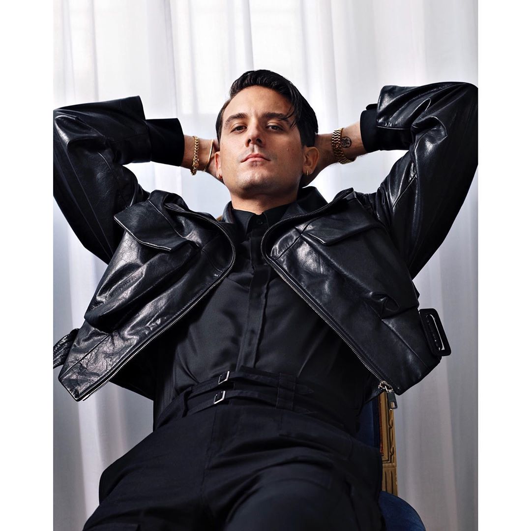 G-Eazy poses in black leather jacket and pants, hands behind his head, neutral expression, seated indoors with curtain backdrop.