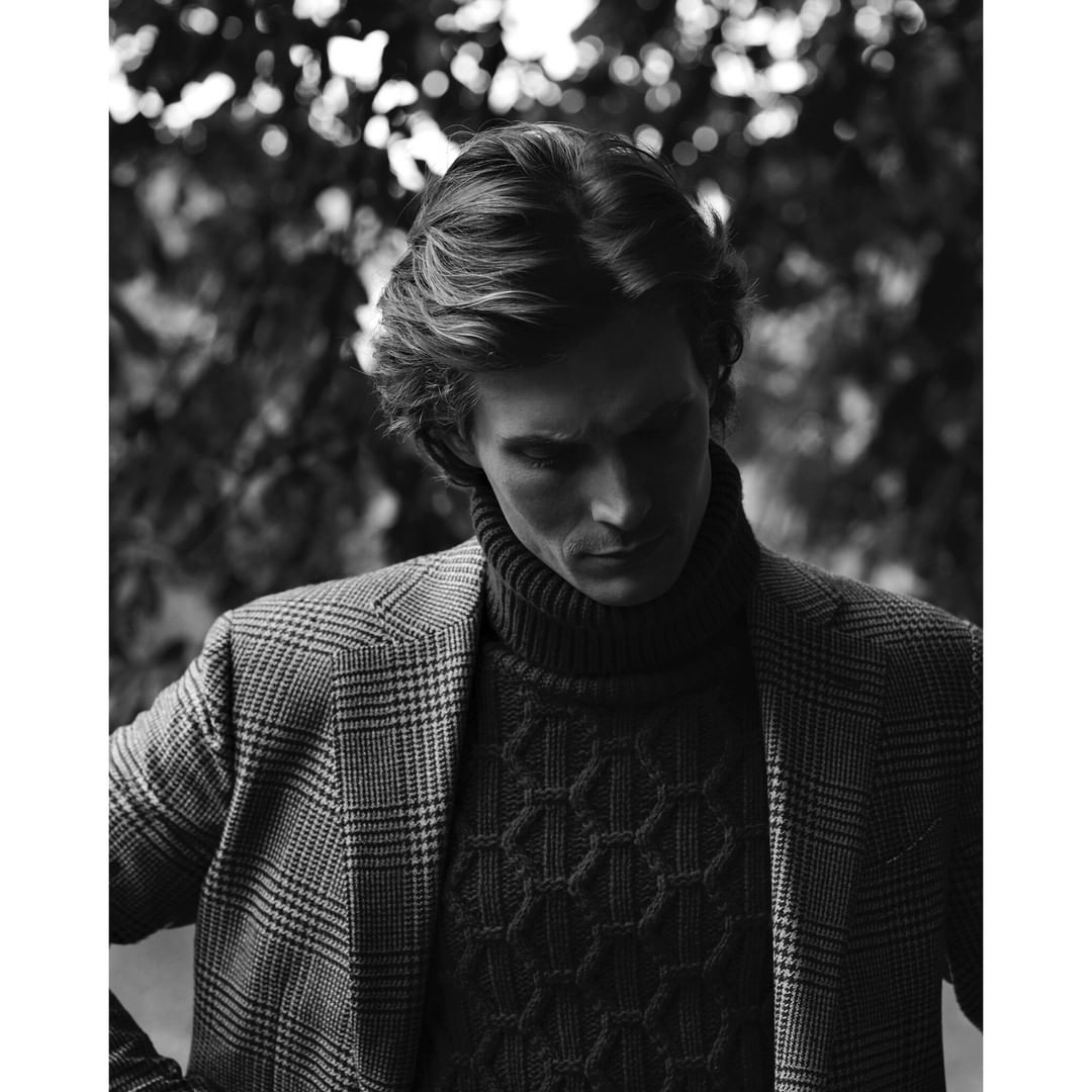 Black and white portrait of a man in a patterned sweater and blazer posing outdoors against a blurred leafy backdrop in natural light.