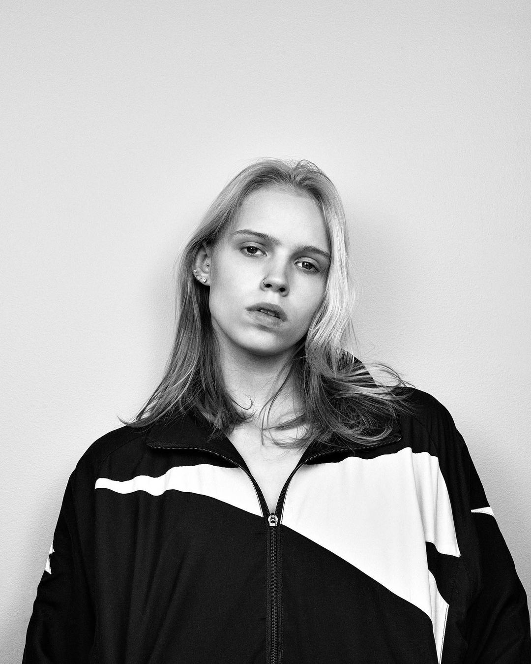 Black and white portrait of a young adult woman with light blonde hair wearing a black and white track jacket against a plain background.