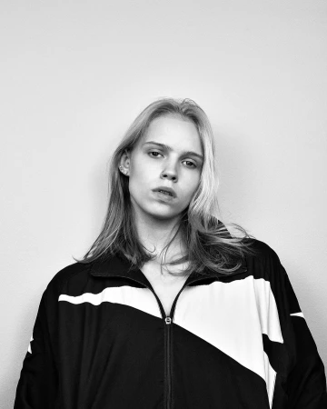 Black and white portrait of a young adult woman with light blonde hair wearing a black and white track jacket against a plain background.