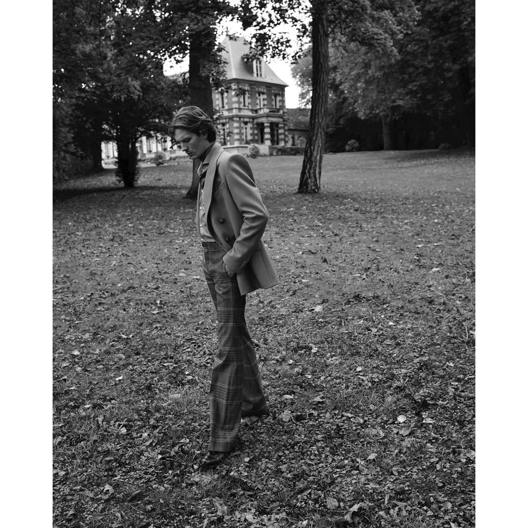 A man stands in a field in front of a large house, wearing a blazer and flared pants, captured in a black and white, medium shot.