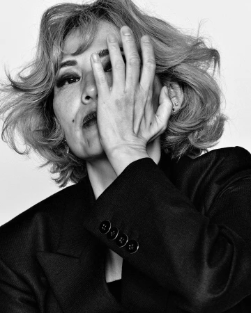 Monochrome portrait of a woman with short wavy hair wearing a dark blazer, holding her hand over her face.