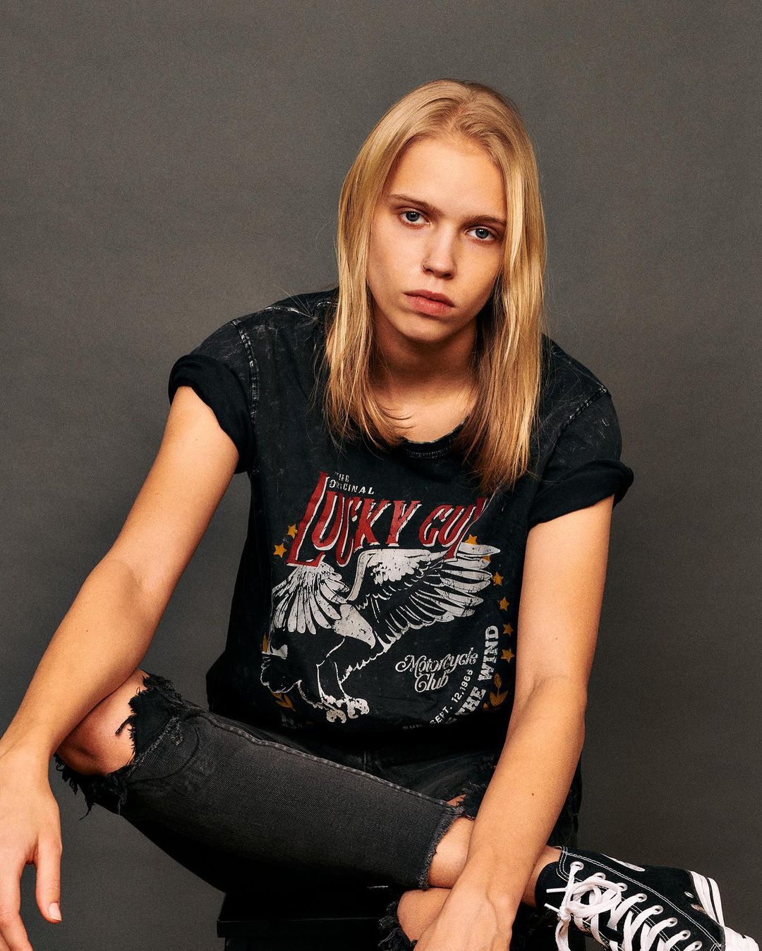 Young adult posing indoors with a graphic tee shirt, ripped black jeans, and black-and-white sneakers against a solid gray backdrop.