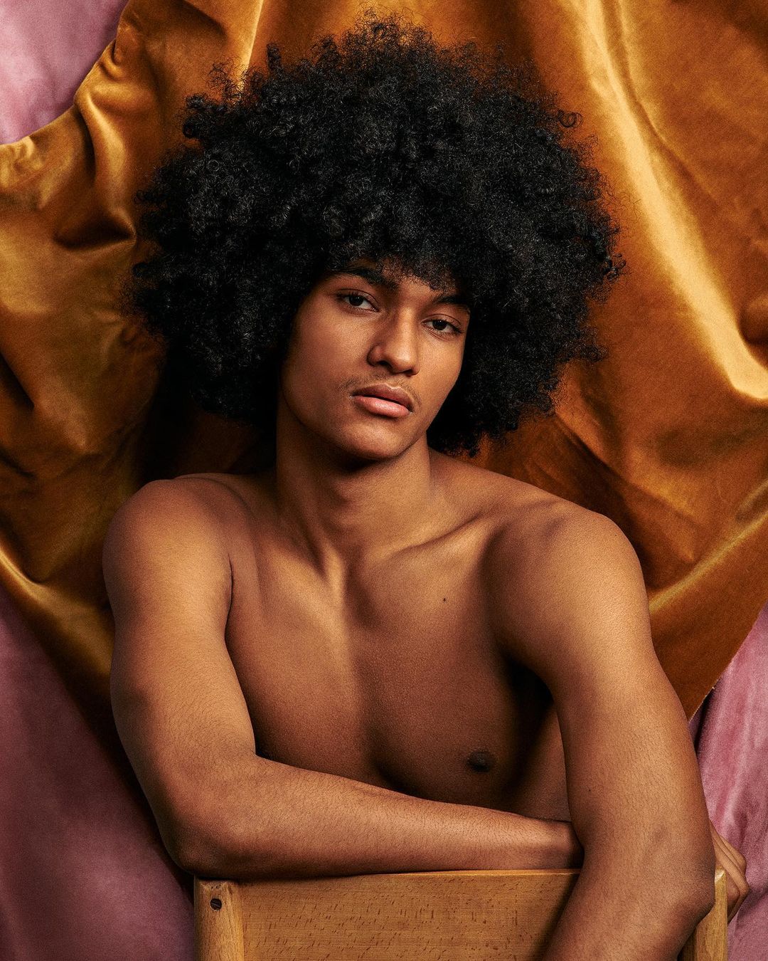 Portrait of young man with large black afro hairstyle resting arms on wooden chair against draped textured fabric background