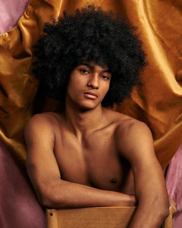 Portrait of young man with large black afro hairstyle resting arms on wooden chair against draped textured fabric background