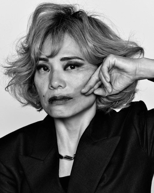 Close-up portrait of a mature adult female with short wavy hair, wearing a blazer and silver necklace in a black and white studio shot.