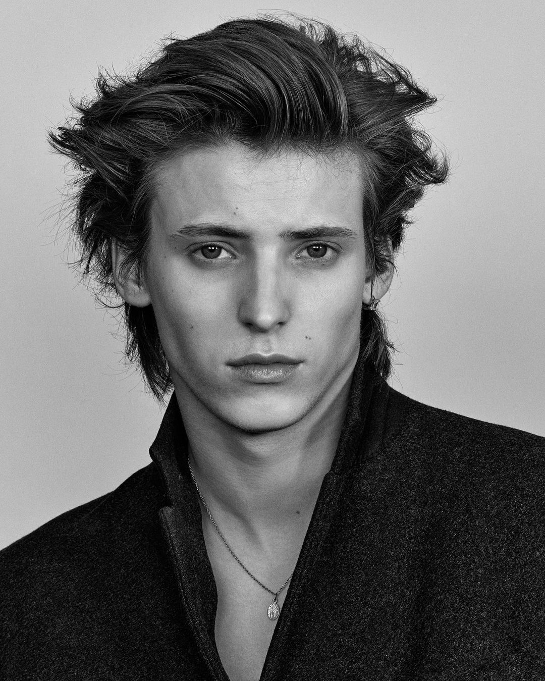 Black and white portrait of an adult male with tousled brown hair wearing a dark collared coat and a pendant necklace looking at the camera.