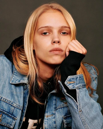 Close-up portrait of a blonde model wearing a denim jacket and black hoodie resting head on hand