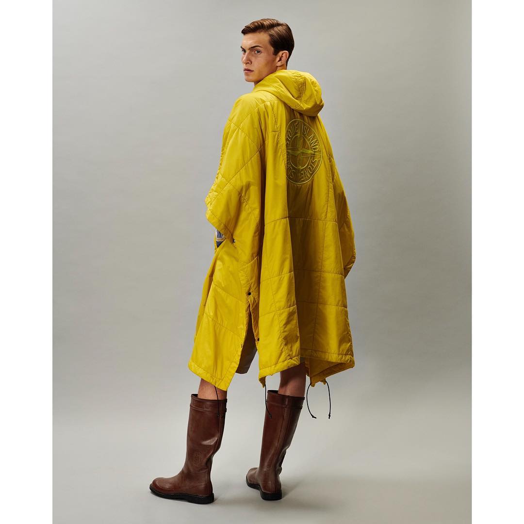 Studio shot of a man in a hooded, quilted yellow poncho and brown boots, looking over his shoulder against a neutral background.
