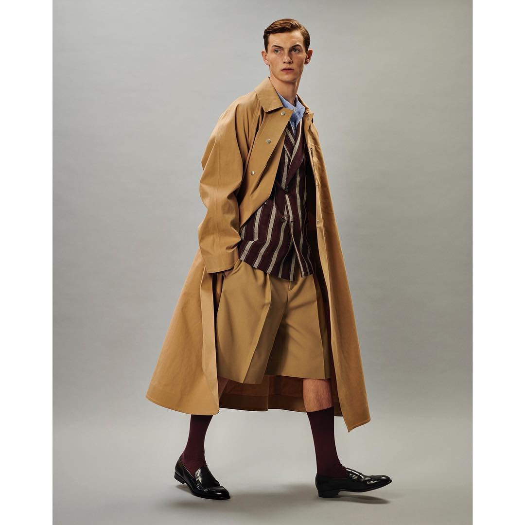 Male fashion model in a tan trench coat, striped vest, and shorts, walking in a studio setting. Classic, stylish men's apparel.