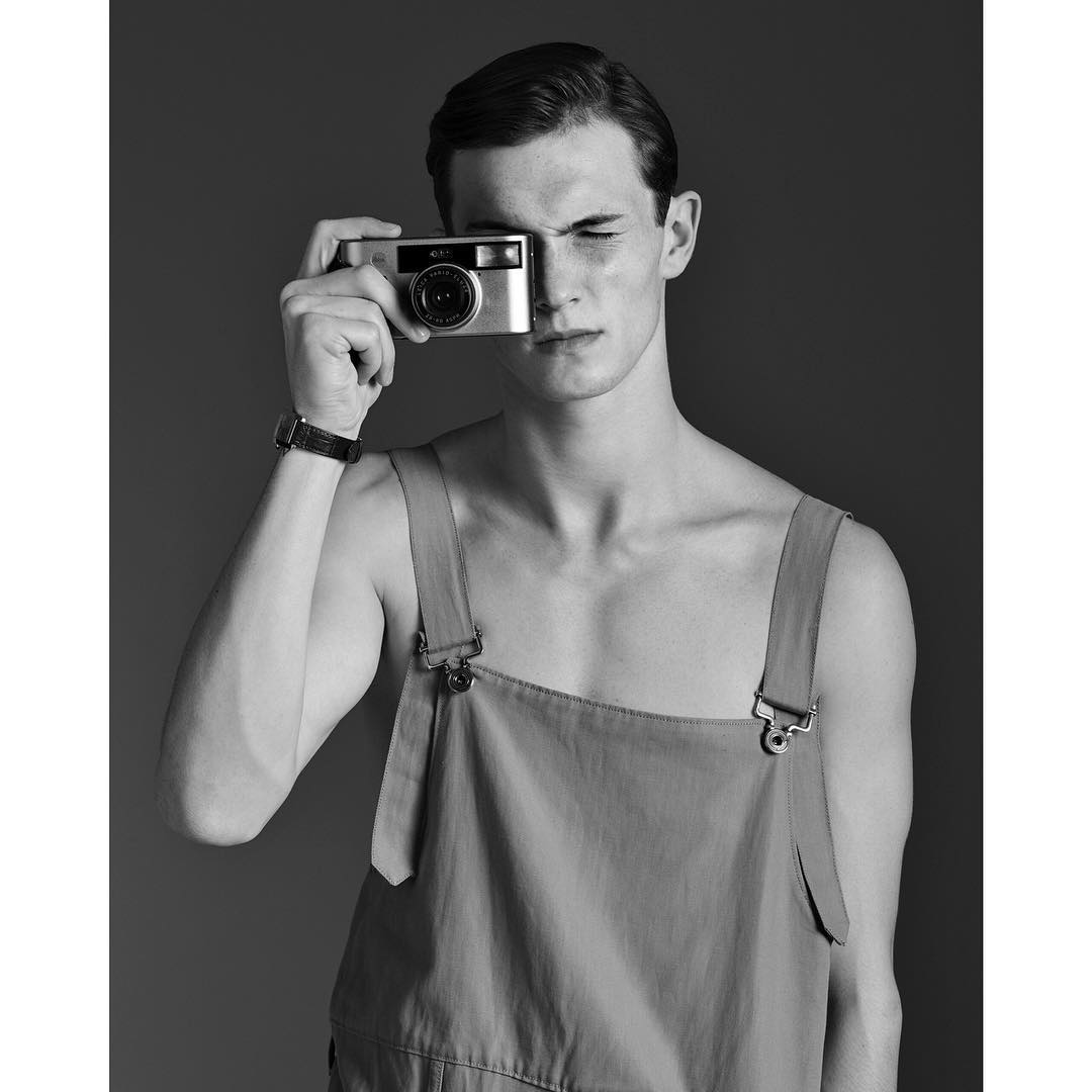 Monochrome portrait of a young adult male model in overalls holding a vintage camera up to his face in a photo studio.