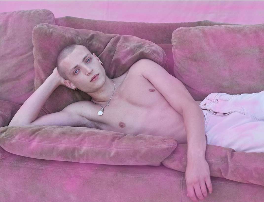 A young, short-haired person lies on a pink couch with white pants and bare torso, leaning back with one hand behind their head.
