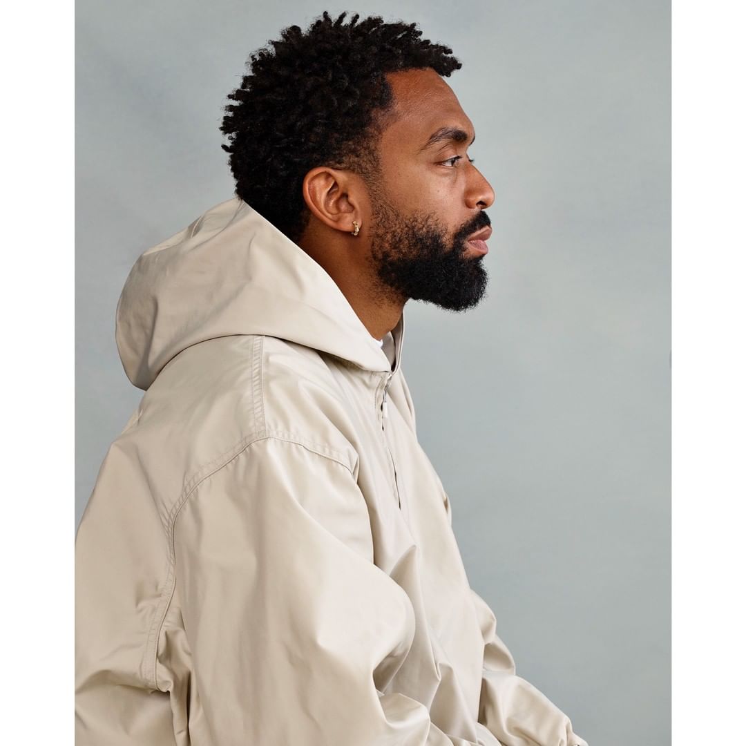 Profile portrait of a Black male with a beard and curly hair wearing a hooded tan jacket against a grey background in a photo studio.
