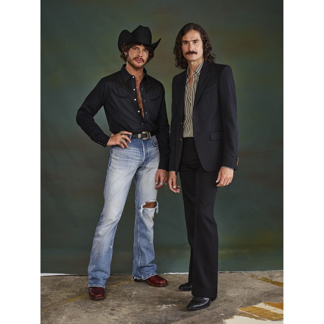 Two men pose in retro fashion: one in a cowboy hat and the other in a blazer, both sporting mustaches, against a muted green backdrop.