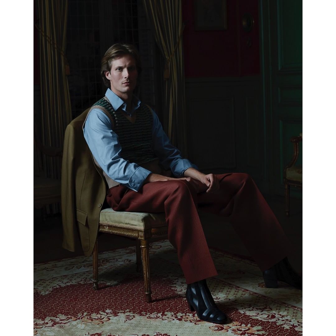 Adult male seated in a room with patterned carpet and a coat draped on the chair, creating a classic and posed fashion portrait.