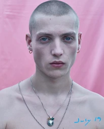 Close-up portrait of a young man with a shaved head, blue eyes, silver hoop earrings, and layered necklaces against a pink background.