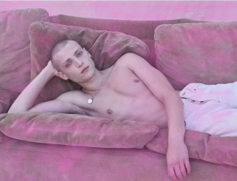 Shirtless man with a buzz cut lounging on a pink couch wearing light pants